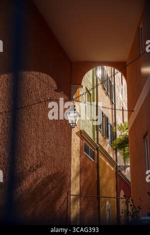 La lumière chaude du soleil illumine une étroite ruelle européenne, soulignant les murs texturés, les fenêtres à volets et une lanterne suspendue sous une arche. Banque D'Images