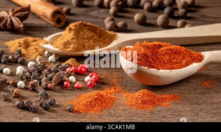 Une image captivante avec deux cuillères en bois remplies d'épices vibrantes comme le paprika et le curcuma, entourées de poivre noir et rose dispersés Banque D'Images