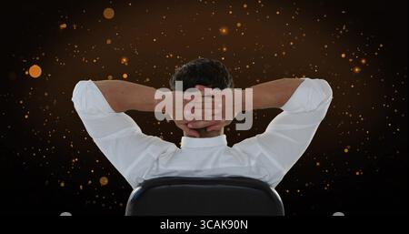 Mâle en chemise blanche serrant les mains derrière la tête en studio, avec chaise de bureau noire, étincelles incandescentes Banque D'Images