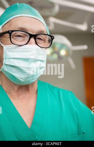 Chirurgien masculin senior portant des gommages verts, une casquette, un masque et des lunettes debout sous OU à la lumière Banque D'Images