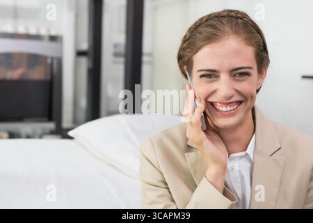 Femme d'affaires portant blazer beige, assise sur le lit parlant sur smartphone dans la chambre d'hôtel, espace copie Banque D'Images