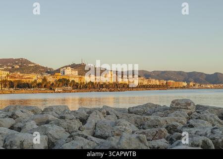 Vue des bâtiments et des montagnes bordant la côte, avec des rochers au premier plan reflétant la lumière du soleil couchant, Salerne, Campanie, Italie. Banque D'Images