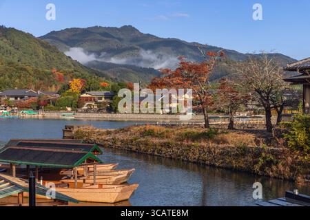 Les bateaux en bois reposent sur la rivière Katsura calme à Arashiyama, Kyoto, Japon, tandis que la brume matinale flotte sur les collines colorées et les maisons traditionnelles le long de la rive tranquille Banque D'Images