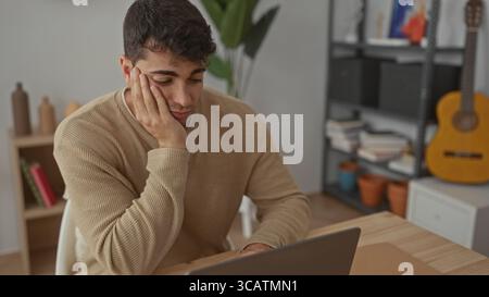 L'homme tient la main pour faire face à bâillement à l'ordinateur portable tandis que le jeune étudiant hispanique travaille au bureau d'ordinateur masculin à la maison. Banque D'Images