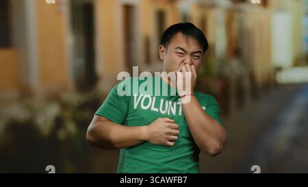 Jeune homme bénévole dans la rue urbaine tenant le nez dans une chemise verte avec expression peinte indiquant l'odeur. Banque D'Images
