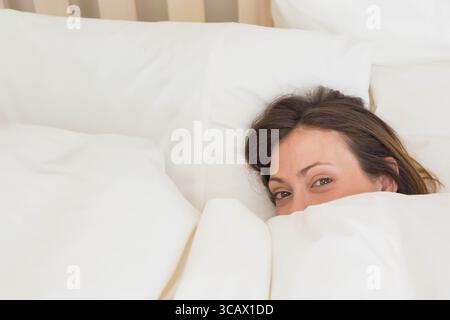 Femme jetant un coup d'œil sous la couette blanche dans la chambre avec des oreillers et des rideaux impeccables, espace de copie Banque D'Images