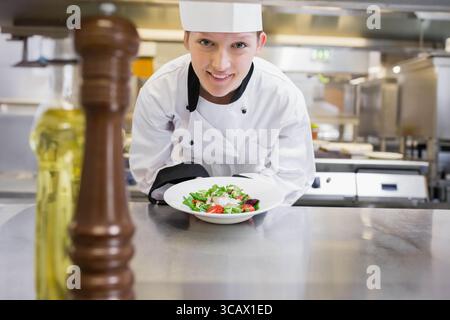 Femme chef en uniforme penchant au comptoir montrant une salade fraîche avec moulin à poivre dans la cuisine Banque D'Images