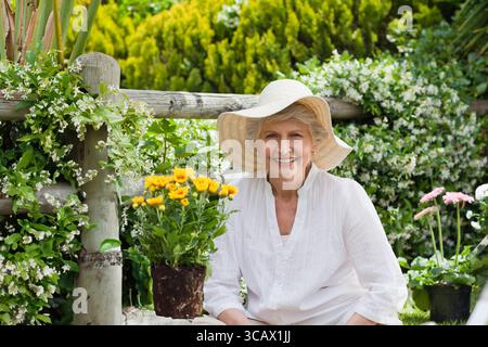 Jardinier senior s'occupant de mamans jaunes et de fleurs roses dans la cour arrière avec chapeau de paille et chemisier blanc Banque D'Images