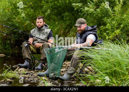 Deux pêcheurs en pleine vitesse assis près d'une rivière de montagne, l'un tenant un filet avec un poisson fraîchement pêché, tandis que l'autre se concentre sur sa canne à pêche, appréciant Banque D'Images