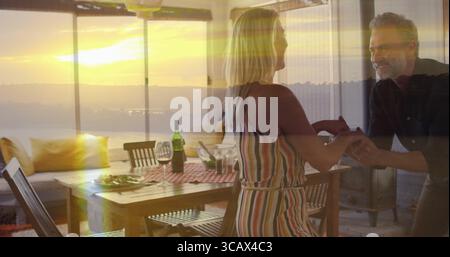 Couple mûr dansant portant une robe rayée, chemise sombre dans la salle à manger au coucher du soleil, avec bouteille de vin Banque D'Images