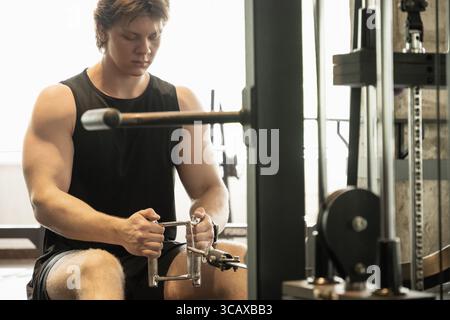 Jeune homme caucasien sportif portant un débardeur sans manches vierge s'exerçant sur une machine à ramer basse au gymnase. Banque D'Images