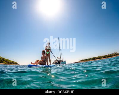Blue Lagoon, Croatie - 9 septembre 2023 : deux femmes profitent d'une journée ensoleillée sur une planche à aubes dans les eaux cristallines, avec un voilier ancré à proximité. Banque D'Images
