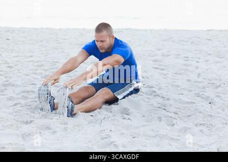 Homme en forme s'étirant vers l'avant tout en atteignant les baskets sur la plage de sable près du rivage Banque D'Images