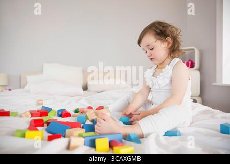 Enfant en bas âge féminin jouant avec des blocs de bois colorés sur un grand lit sous la lumière douce de la fenêtre Banque D'Images