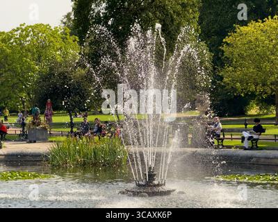 La lumière du soleil scintille sur l'eau depuis une fontaine dans les jardins italiens, avec des gens se relaxant sur des bancs en arrière-plan, Hyde Park, Londres, Royaume-Uni. Banque D'Images