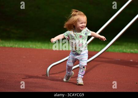 Un tout-petit aux cheveux rouges qui court joyeusement sur une surface de jeu en caoutchouc, entouré d'herbe verte. L'enfant est énergique et habillé dans le jeu coloré CLO Banque D'Images