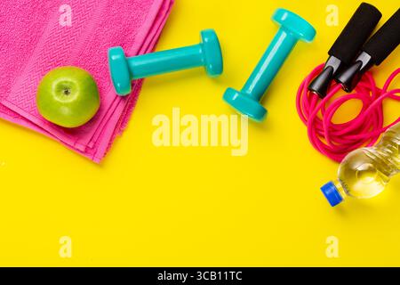 Concept fitness et sport. Corde à sauter, pomme verte, haltères, bouteille d'eau et serviette rose sur fond jaune vif. Espace libre. Banque D'Images