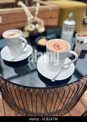 Deux tasses blanches de cappuccino surmontées de mousse et de cannelle, servies sur des soucoupes sur une table ronde en métal noir. Banque D'Images