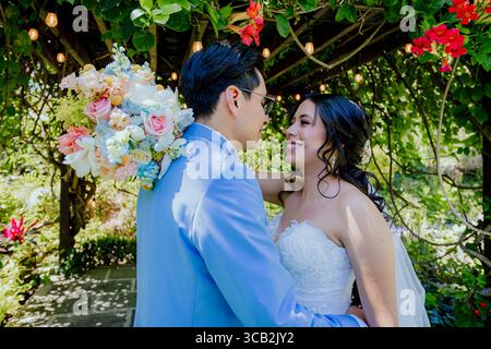 Le marié en costume bleu clair tient un bouquet de fleurs pastel tout en embrassant sa mariée dans une élégante robe en dentelle sans bretelles, sous une pergola en bois décorée Banque D'Images