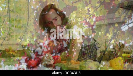 Homme barbu portant tablier tranchant pomme avec couteau à table de cuisine, avec tablette et feuilles d'automne Banque D'Images