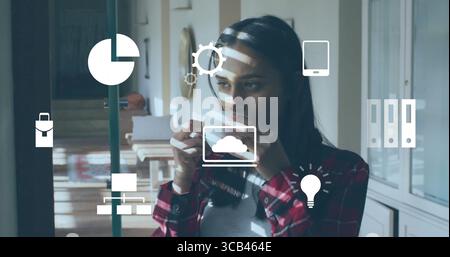 Femme asiatique buvant portant une chemise rouge à carreaux tenant une tasse en céramique dans le bureau à domicile, avec des icônes technologiques Banque D'Images