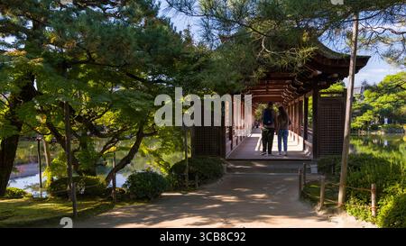 Deux personnes marchent main dans la main le long d'un pavillon traditionnel Heian Jingu Taiheikaku entouré d'une verdure naturelle pittoresque et d'un soleil éclatant. Le settin Banque D'Images