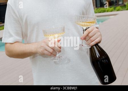 Homme en chemise blanche tenant deux verres de coupe de champagne et une bouteille sombre lors d'une célébration en plein air Banque D'Images