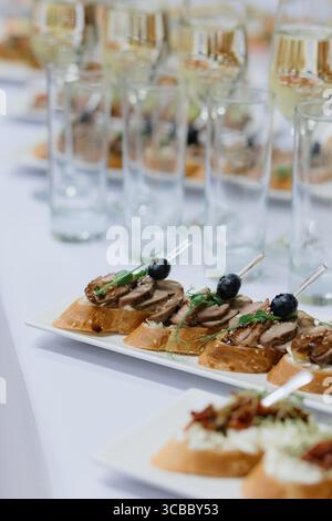 Canapés gourmands avec de la viande tranchée, des bleuets et des herbes sur des tranches de pain servies sur une table blanche avec des verres de vin mousseux Banque D'Images