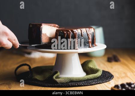 Tranchant à la main un riche cheesecake à la croûte foncée, avec des couches crémeuses de vanille et de chocolat, surmonté de ganache au chocolat brillant. Banque D'Images