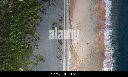 Polynésie française, île de Rangiroa : vue aérienne capturant une mince route séparant la végétation luxuriante des palmiers verts de la plage de sable et des vagues de l'océan. Vol drone de haut en bas Banque D'Images