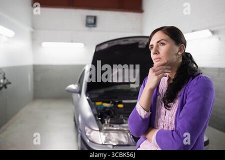 Femme debout dans le garage inspectant le moteur à côté de la berline argentée sous des lumières fluorescentes, espace de copie. Automobile, réparation, entretien, industriel, diag Banque D'Images