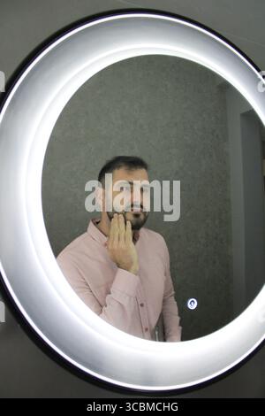 Homme en chemise rose regardant son reflet dans un miroir lumineux rond, touchant son visage avec soin. Banque D'Images