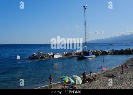 Messine, Sicile, Italie – août 2025. Le vice-premier ministre et ministre de l’infrastructure, Matteo Salvini, assiste à un déjeuner au restaurant Grecale pour célébrer l’approbation par le CIPESS du projet controversé du pont du détroit de Messine. Sur la plage voisine, les baigneurs partagent le sable avec les manifestants tenant un drapeau « No Ponte » (No Bridge) contre la construction. Dans l'eau, un bateau de pêche traditionnel de Messine appelé felouca, utilisé pour la chasse à l'espadon, est décoré de drapeaux italiens de la Lega Nord de droite. Villa San Giovanni en Calabre est visible en arrière-plan. Banque D'Images