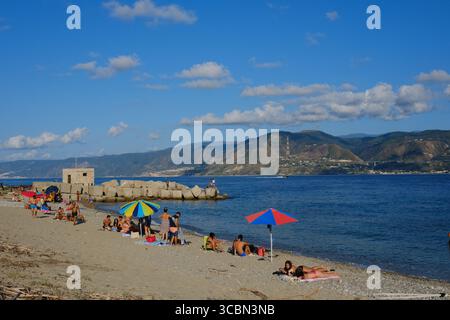 Messine, Sicile, Italie – août 2025. Le vice-premier ministre et ministre de l’infrastructure, Matteo Salvini, assiste à un déjeuner au restaurant Grecale pour célébrer l’approbation par le CIPESS du projet controversé du pont du détroit de Messine. Sur la plage voisine, les baigneurs partagent le sable avec les manifestants tenant un drapeau « No Ponte » (No Bridge) contre la construction. Dans l'eau, un bateau de pêche traditionnel de Messine appelé felouca, utilisé pour la chasse à l'espadon, est décoré de drapeaux italiens de la Lega Nord de droite. Villa San Giovanni en Calabre est visible en arrière-plan. Banque D'Images