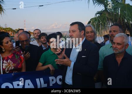 Messine, Sicile, Italie – août 2025. Le vice-premier ministre et ministre de l’infrastructure, Matteo Salvini, assiste à un déjeuner au restaurant Grecale pour célébrer l’approbation par le CIPESS du projet controversé du pont du détroit de Messine. Sur la plage voisine, les baigneurs partagent le sable avec les manifestants tenant un drapeau « No Ponte » (No Bridge) contre la construction. Dans l'eau, un bateau de pêche traditionnel de Messine appelé felouca, utilisé pour la chasse à l'espadon, est décoré de drapeaux italiens de la Lega Nord de droite. Villa San Giovanni en Calabre est visible en arrière-plan. Banque D'Images