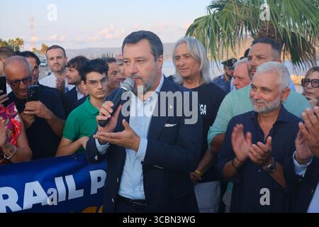 Messine, Sicile, Italie – août 2025. Le vice-premier ministre et ministre de l’infrastructure, Matteo Salvini, assiste à un déjeuner au restaurant Grecale pour célébrer l’approbation par le CIPESS du projet controversé du pont du détroit de Messine. Sur la plage voisine, les baigneurs partagent le sable avec les manifestants tenant un drapeau « No Ponte » (No Bridge) contre la construction. Dans l'eau, un bateau de pêche traditionnel de Messine appelé felouca, utilisé pour la chasse à l'espadon, est décoré de drapeaux italiens de la Lega Nord de droite. Villa San Giovanni en Calabre est visible en arrière-plan. Banque D'Images