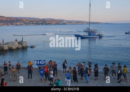 Messine, Sicile, Italie – août 2025. Le vice-premier ministre et ministre de l’infrastructure, Matteo Salvini, assiste à un déjeuner au restaurant Grecale pour célébrer l’approbation par le CIPESS du projet controversé du pont du détroit de Messine. Sur la plage voisine, les baigneurs partagent le sable avec les manifestants tenant un drapeau « No Ponte » (No Bridge) contre la construction. Dans l'eau, un bateau de pêche traditionnel de Messine appelé felouca, utilisé pour la chasse à l'espadon, est décoré de drapeaux italiens de la Lega Nord de droite. Villa San Giovanni en Calabre est visible en arrière-plan. Banque D'Images