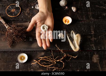 Main féminine avec coquille entourée de bougies, racines, mèches de cheveux et objets magiques sur la surface en bois comme un rituel de sorcière Banque D'Images