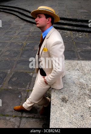 Un homme portant un chapeau de paille et un costume de couleur claire est appuyé contre une structure en pierre, avec une main dans sa poche. Banque D'Images