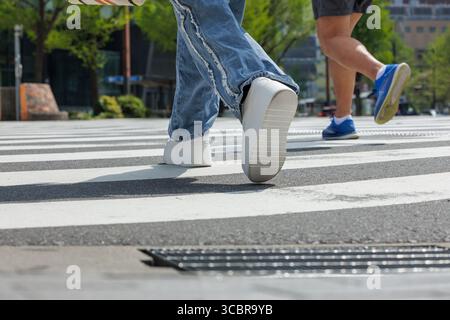 L'angle bas de deux personnes traversant l'intersection montre le mouvement. Capture de mi-foulée, met en valeur des tenues urbaines décontractées - jeans bleus et vêtements de course à pied. Banque D'Images