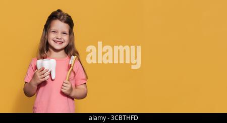 Une fille heureuse tient une brosse à dents et une dent en plastique dans ses mains. Un cliché panoramique isolé. Un endroit pour votre texte. prévention de la carie infantile. Banque D'Images