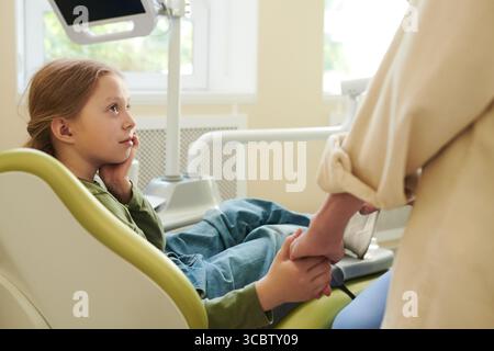Fille d'enfant caucasienne assise dans la chaise dentaire tenant la main de la femme adulte, regardant vers le haut avec une expression réfléchie, équipement dentaire visible en arrière-plan, recevant le réconfort Banque D'Images