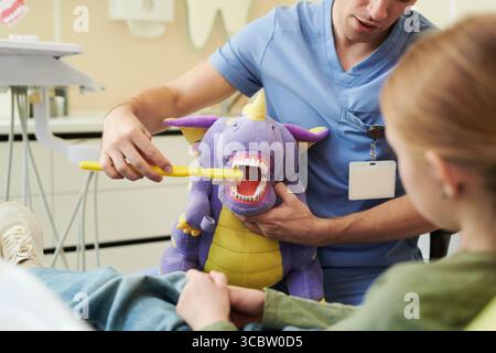 Dentiste masculin caucasien démontrant la technique de brossage des dents à l'aide d'une grande brosse à dents et d'une marionnette de dragon violette à une fille caucasienne, assise dans un fauteuil dentaire pendant une session d'éducation dentaire Banque D'Images