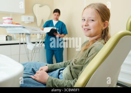 Fille caucasienne assise dans le fauteuil dentaire souriant à la caméra, tandis que dentiste féminine dans les gommages debout en arrière-plan lisant le dossier patient dans le cadre de clinique dentaire moderne Banque D'Images