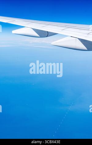 Vue en vol de l'aile blanche d'un airbus A350-900, volant au-dessus de la mer du Nord avec une rangée d'éoliennes dans la mer. Ciel bleu profond, petit nuage. Banque D'Images