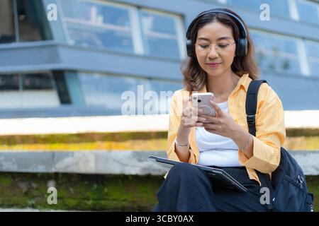 Une femme est assise sur un rebord avec son téléphone et son ordinateur portable. Elle porte des écouteurs et elle est concentrée sur son téléphone Banque D'Images