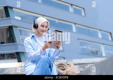 Une femme portant un foulard est assise sur un rebord avec une tablette à la main. Elle est souriante et elle profite de son temps Banque D'Images