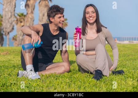 Couple se reposant sur l'herbe avec des bouteilles d'eau après l'entraînement en plein air sur une journée ensoleillée Banque D'Images