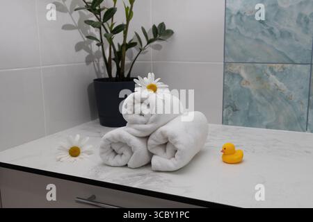 Serviettes blanches douces sont soigneusement roulées et empilées sur un comptoir en marbre, accompagnées de fleurs fraîches et d'un canard en caoutchouc mignon, créant une atmosphère calme et accueillante dans la salle de bain. Banque D'Images