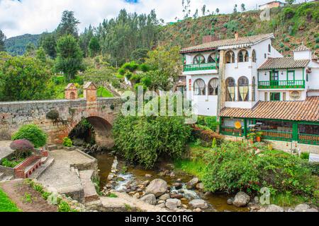 Vue panoramique sur les bâtiments coloniaux et un pont pavé à Monguí, Boyacá, mettant en valeur une végétation luxuriante et des eaux sereines. Banque D'Images